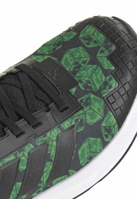 Chaussure de sport noire avec une tige en tissu texturé ornée de motifs verts en blocs, des accents moulés, et une semelle en caoutchouc blanche.