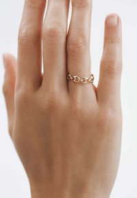 Anillo de cadena de oro en un dedo esbelto, con eslabones ovalados entrelazados y un acabado liso y pulido. La piel tiene un tono claro y una textura sutil.