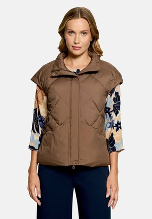 Femme portant une veste matelassée marron sans manches par-dessus une chemise fleurie à manches longues et un pantalon foncé, debout devant un fond uni.