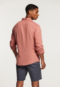 Chemise pour homme de couleur rose clair, manches longues retroussées, détails au col, accompagnée d'un short gris foncé. Le tissu semble texturé et respirant.