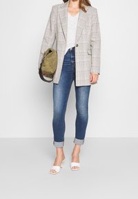 Blazer oversize à carreaux beige et noir, haut gris, jean bleu slim et sandales à talons blancs. Comprend des manches retroussées et un sac vert.
