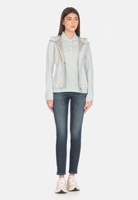Giacca leggera blu chiaro con cappuccio, zip dorata, abbinata a una polo blu chiaro e jeans scuri. Sneakers bianche e rosa completano il look.