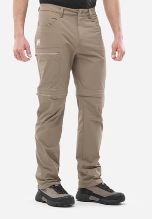 Homme portant un pantalon de randonnée convertible beige avec jambes amovibles zippées et chaussures de randonnée noires et grises sur fond blanc.