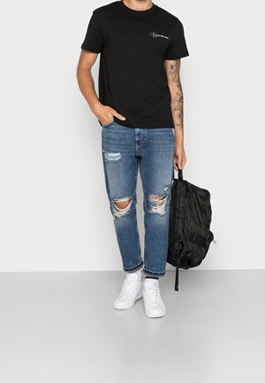 Black cotton t-shirt with logo, ripped blue denim jeans, white sneakers, and a black backpack. Casual outfit with a relaxed fit and distressed details.