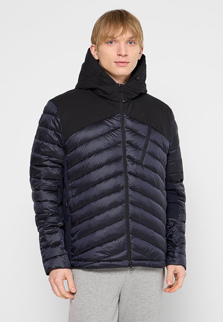 Junger Mann in einer schwarzen und marineblauen gesteppte Kapuzenjacke mit Reißverschlusstasche auf der Brust und hellgrauen Hosen, stehend vor einem schlichten Hintergrund.