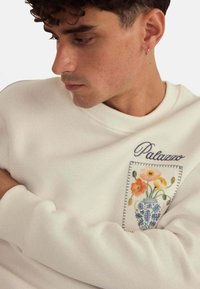 Sweat-shirt blanc avec des poignets côtelés, orné d'une broderie florale sur un patch. Le design comprend un vase avec des fleurs colorées et des accents bleus.