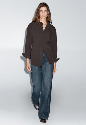 Zwarte button-up shirt met een relaxte pasvorm en unieke draping, gecombineerd met hoog-waist, wijde denimbroek in een donkere tint en bruine platte schoenen.