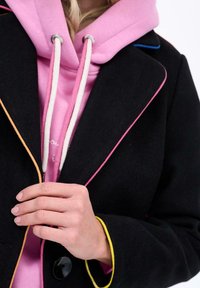 Osoba v ružovej mikine s bielymi šnúrkami a čiernom kabáte s viacfarebnými pruhmi, ktorá drží klopu kabátu.