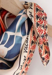 Écharpe en soie avec motif géométrique bleu et bordure florale rouge, drapée sur une tige en bois, avec un texte noir en cursive le long du bord.