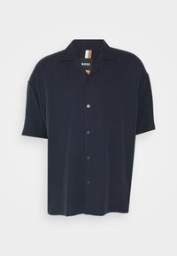 Camisa de manga corta abotonada en azul oscuro, con cuello, botones frontales y una textura suave. Etiqueta visible en el cuello con rayas en contraste.