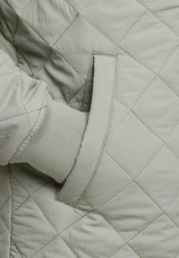 Gepolsterte, hellgraue Jacke mit gerippten Bündchen und Seitentaschen, die eine weiche Textur und ein diagonales Steppmuster aufweist.