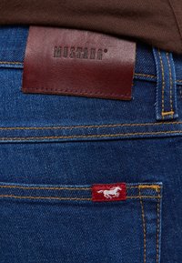 Granatowe dżinsy z ciemnego denimu z brązową skórzaną metką w talii z wygrawerowanym napisem "MUSTANG" oraz czerwoną naszywką z białym logo konia.