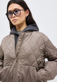 Νεαρή γυναίκα που φοράει μεγάλα τετράγωνα γυαλιά ηλίου, μια παλαιού χρώματος μπουφάν με το φερμουάρ κλειστό και κάτω από αυτό μια σκούρα γκρι κουκούλα.