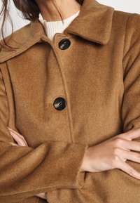 Brun uldfrakke med krave, udstyret med sorte knapper og en blød, struktureret overflade. Stoffet ser varmt og hyggeligt ud.