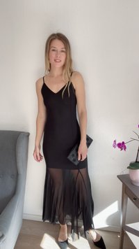 Vestido preto com corpete justo e bainha transparente com franjas. Combinado com uma carteira preta e sapatos de salto cinzas. Em frente a uma parede e mobiliário claros.