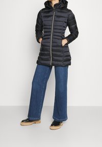 Sort pufferjakke med hætte, der har lodret quiltet design, frontlynlås og sidelommer. Kombinationen med brede blå jeans og sorte sko.