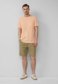 Hellbeige T-Shirt mit einer Brusttasche, kombiniert mit olivgrünen Shorts. Die Schuhe sind cremefarben mit hellbraunen Akzenten. Glatte Stofftextur.