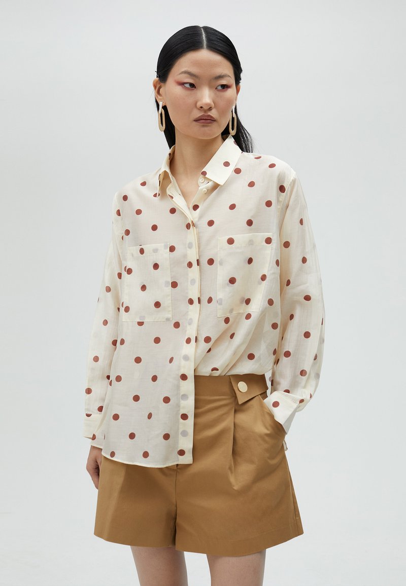 Cream blouse with large brown polka dots, two chest pockets, and button-up design, paired with light brown, high-waisted shorts.