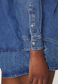 Lähikuva tummaihoisesta kädestä, joka lepää sivulla, ja jossa on päällään sininen farkkupaita, jossa on pitkät hihat, sekä siihen sopiva hame hopeisilla logopainikkeilla.