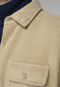 Beżowa, miękka kurtka polarowa z kołnierzem i kieszenią na piersi z srebrnym guzikiem. Pod spodem ciemny prążkowany golf.