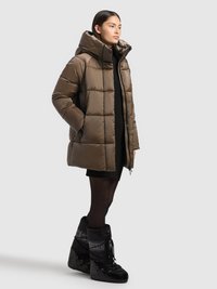 Brūna polsterēta pūkaina jaka ar augstu apkakli, vītņotu dizainu un sānu kabatām. Valda ar melniem ceļgalu augstiem ziemas zābakiem un melnu kleitu.