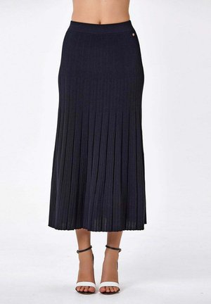 Femme portant une jupe midi noire plissée taille haute, associée à des talons blancs à bout ouvert avec bride à la cheville.