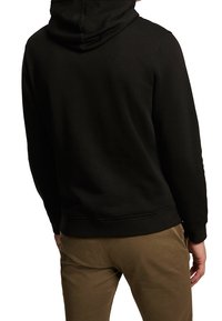 Personne portant un sweat à capuche noir et un pantalon marron, vue de dos avec une main détendue sur le côté.