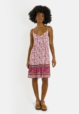 Mujer con peinado afro que lleva un vestido sin mangas floral de color rosa y púrpura hasta la rodilla y sandalias marrones de plataforma, posando sobre fondo blanco.