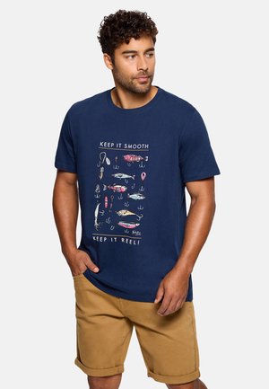 Hombre con camiseta azul marino con gráfico de señuelos y anzuelos de pesca coloridos y pantalones cortos color caqui, de pie contra un fondo blanco.