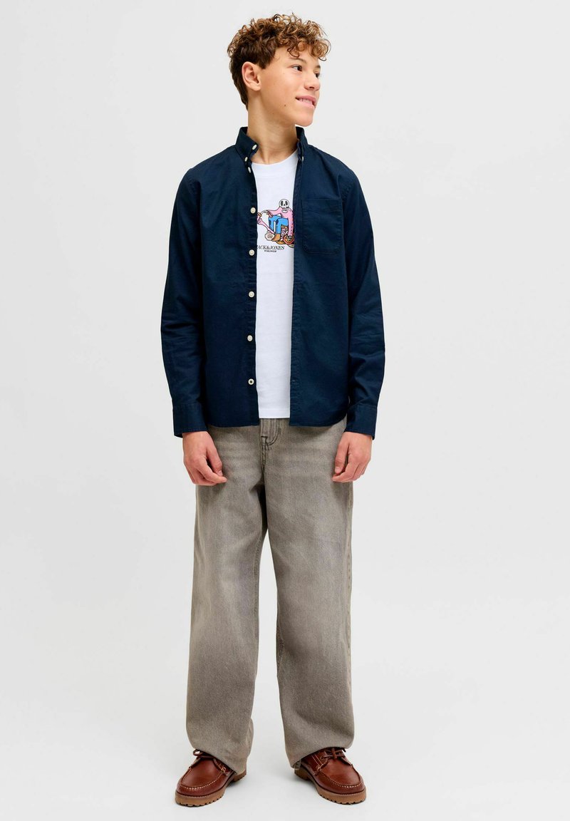 Uue korteraga teismeline poiss kannab tumesinist lahtist särki valge graafilise T-särgi peal, lahtised beežid püksid ja pruunid nahast kingad, seisab ja vaatab paremale.