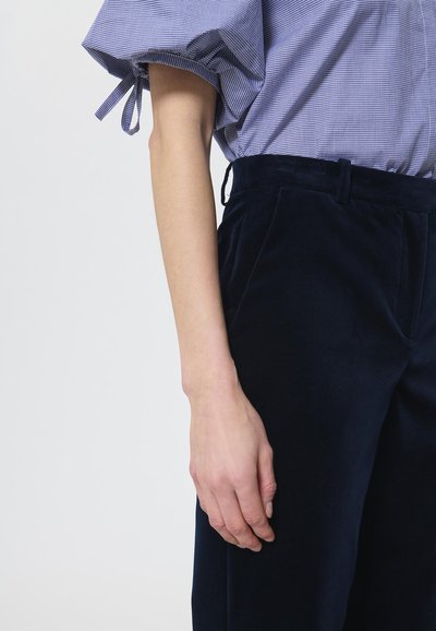 Tamsiai mėlynos aksominės kelnės su lygiu paviršiumi, turinčios priekines kišenes ir pritaikytą siluetą, derinamos su šviesiai mėlynu languotu bluzonu.