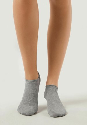 Zwei nackte Beine mit niedrig geschnittenen, grauen Knöchelsocken, die vor einem schlichten, hellen Hintergrund stehen.