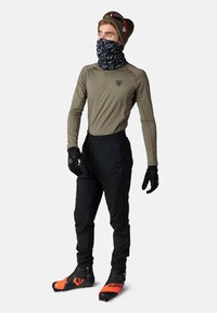 Technisches Outdoor-Outfit mit einem leichten olivfarbenen Langarmshirt, schwarzen Hosen, schwarzen Handschuhen und orange akzentuierten Stiefeln, ergänzt durch einen gemusterten Halswärmer.