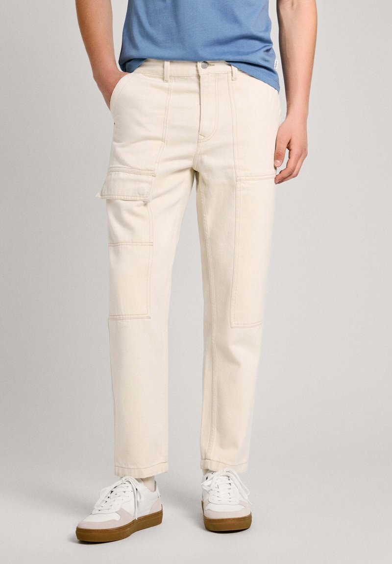 Beige cargo bukser med flere lommer, lige snit og synlige syninger, parret med hvide sneakers og en blå t-shirt.
