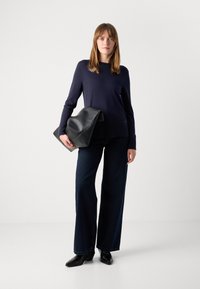 Marineblauwe gebreide trui, wijde donkerblauwe spijkerbroeken en een zwarte leren oversized clutch. Model draagt puntige zwarte enkelboots. Eenvoudig, modern ontwerp.