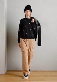 Felpa nera con ricami, pantaloni cargo beige, berretto nero e una giacca di pelle nera appoggiata su una spalla. Sneakers bianche.