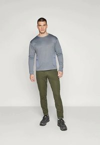 Άνδρας στέκεται φορώντας μακρυμάνικη γκρι μπλούζα, ελαιόχρωμο παντελόνι και μαύρα παπούτσια πεζοπορίας μπροστά από ένα απλό φωτεινό φόντο.