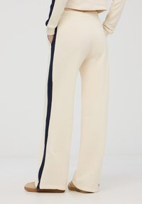 Pantaloni sportivi color crema con una sottile striscia laterale blu navy, tessuto morbido e vita elastica con tasche, indossati con scarpe color beige chiaro.