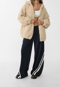 Beige fluffy hooded jacket with zip closure, worn over a white crop top. Paired with navy wide-leg pants featuring white side stripes.
