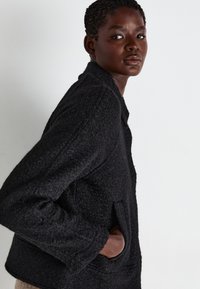Black textured jacket with wide sleeves, a collar, and front pockets, shown from a side angle against a plain background.
