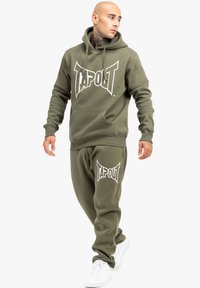 Olīvu zaļš kokvilnas kapucis un sporta bikses komplekts, kas izceļas ar lielu baltu "TAPOUT" logotipu, kengura kabatu un regulējamu auklu jostu.