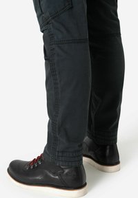 Schwarze Lederschuhe mit roten Schnürsenkeln, kombiniert mit dunkelgrünen Cargo-Hosen, die Seiten Taschen und sichtbare Nähte entlang der Nähte aufweisen.