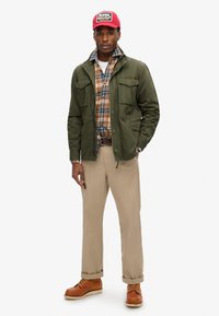 Oliwkowa kurtka z wieloma przednimi kieszeniami noszona na koszuli w kratę. Beżowe spodnie i brązowe buty na sznurowadła dopełniają outfit. Czerwona czapka z logo.