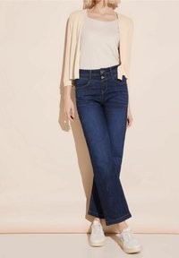 Losse donkeblauwe spijkerbroek met hoge taille en lichte stiksels, gecombineerd met een crèmekleurig kortarmig vest en witte sneakers.