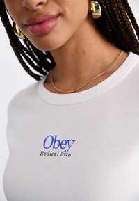 Balts ribots krekls ar zilu druku "Obey" un melnu tekstu "Radical love" zem tā. Valkā zelta ķēdi un auskari ar gredzeniem.