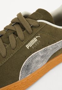 Olivově zelené semišové tenisky s béžovými akcenty, texturovaným šedým logem Puma a hnědou gumovou podrážkou. Vyznačují se silnými tkaničkami a měkkým podšíváním.