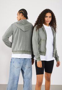 Felpa cropped grigia chiara con zip frontale, tasche frontali e cappuccio. Abbinata a una t-shirt bianca e pantaloni di jeans, indossata da due persone.