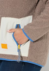 Fleecejacke in Braun, mit einer cremefarbenen Tasche, die einen blauen Reißverschluss und ein oranges Etikett hat. Die Ärmelbündchen haben blaue Akzente; das Material hat eine sichtbare Struktur.