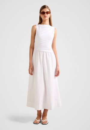 Mujer con vestido midi blanco sin mangas, gafas de sol marrones, pendientes dorados en forma de aro, brazalete y sandalias planas blancas, de pie sobre un fondo liso.