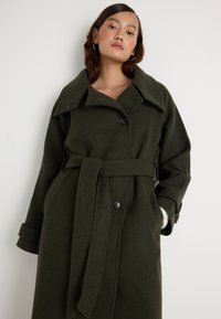 Mulher vestindo um longo casaco de lã verde escuro, com cinto, colarinho largo e botões, de pé contra um fundo branco liso.